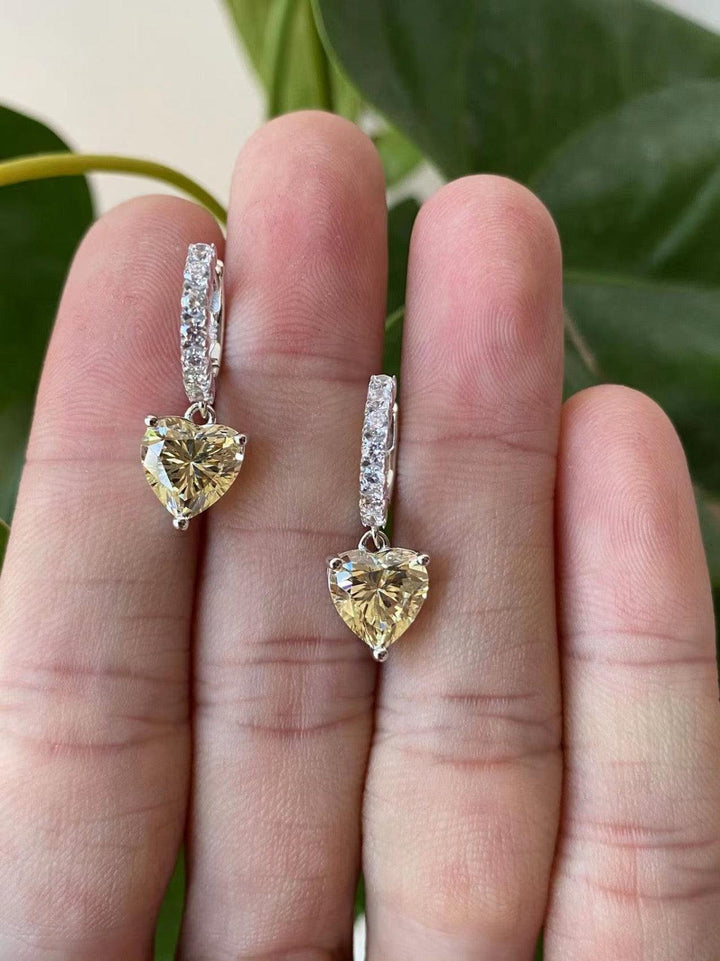A pair of crystal and amber drop earrings held between fingers, with the amber teardrop stones hanging below silver-toned metal settings and clear crystal accents.