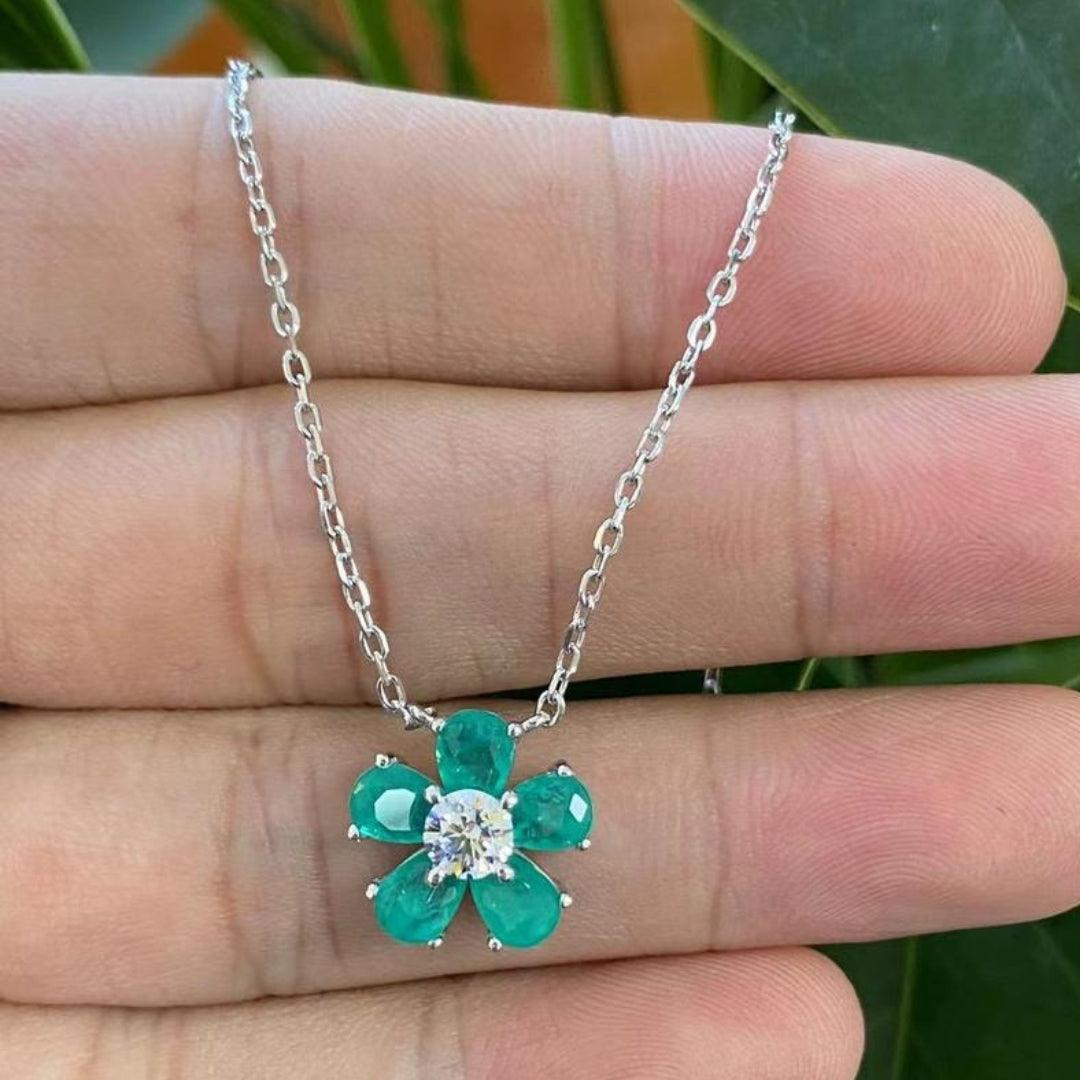 A hand holding a delicate flower-shaped necklace with emerald green crystal petals and a clear crystal center.