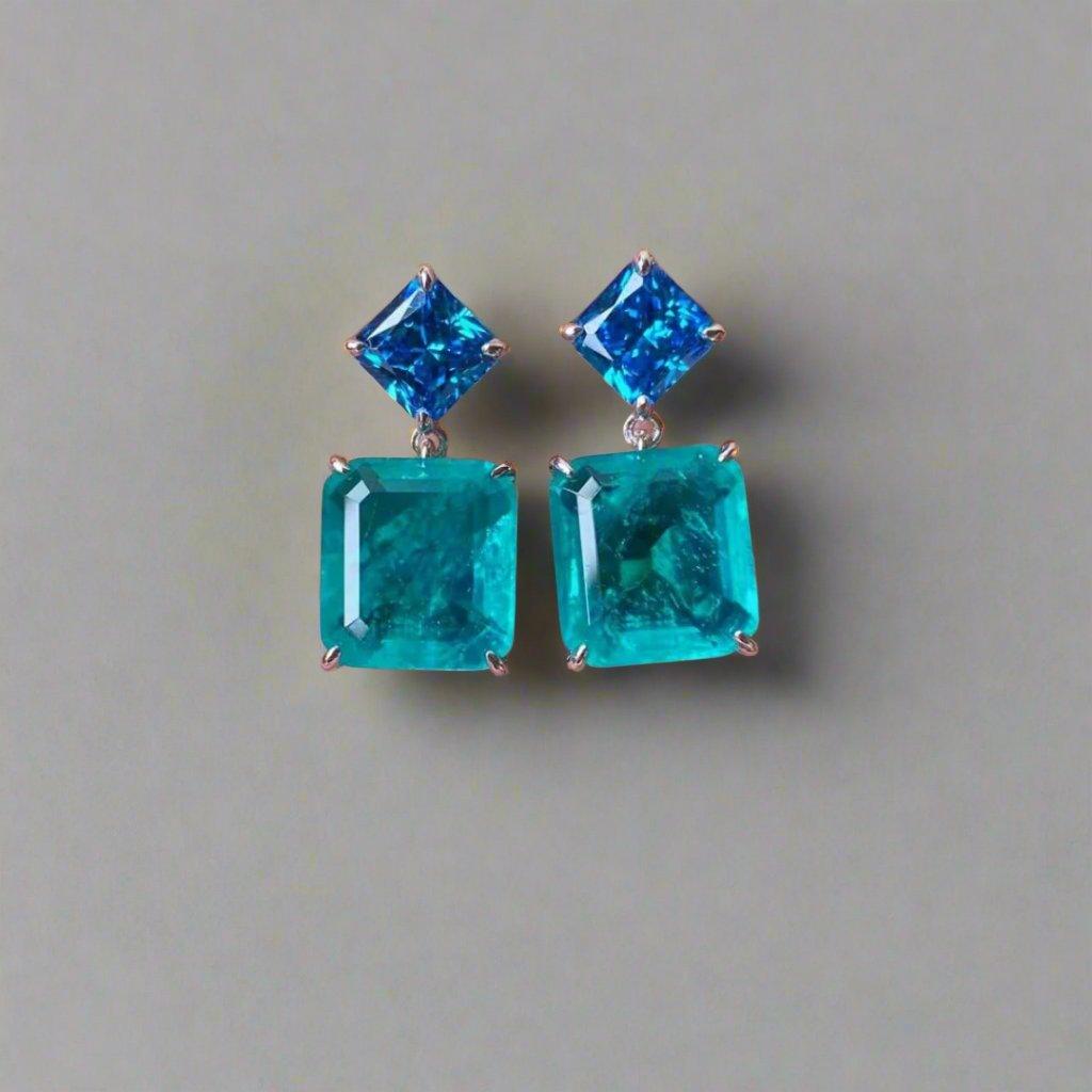A pair of dual-tone blue crystal drop earrings held in a hand, with electric blue square crystal tops and aqua blue rectangular crystal drops, featuring a bright silver metal finish.