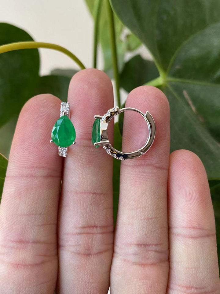 A pair of emerald green teardrop huggie earrings with clear crystal accents, held between fingers against a leafy background.